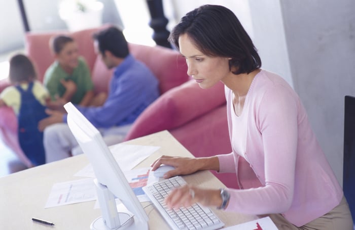 A woman works from home on her computer, with her family in the background.