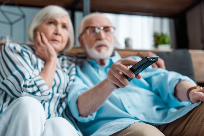 An older couple seem worried as they watch TV on their couch. 