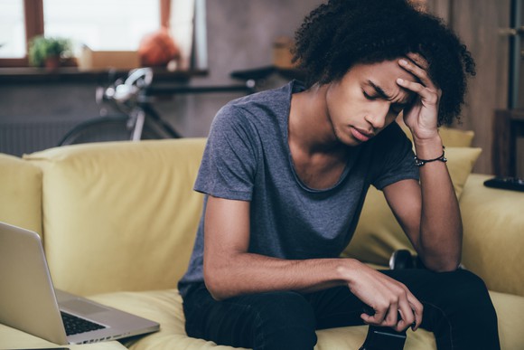 A teenager holds their face in one hand and a video game controller in the other.