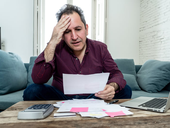 Older man holding a document while holding his head; on the table in front of him are documents, a laptop, and a calculator