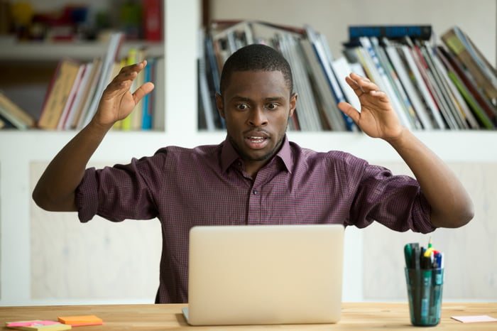 A young man is staring at his laptop in surprise, his arms held up.