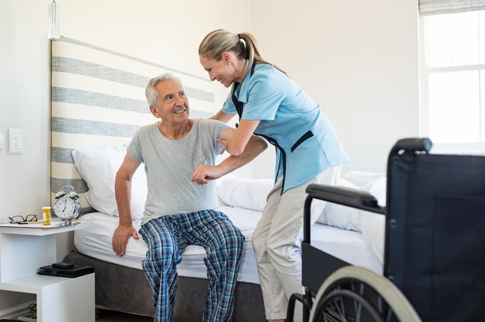 Woman in scrubs helping an older man out of bed