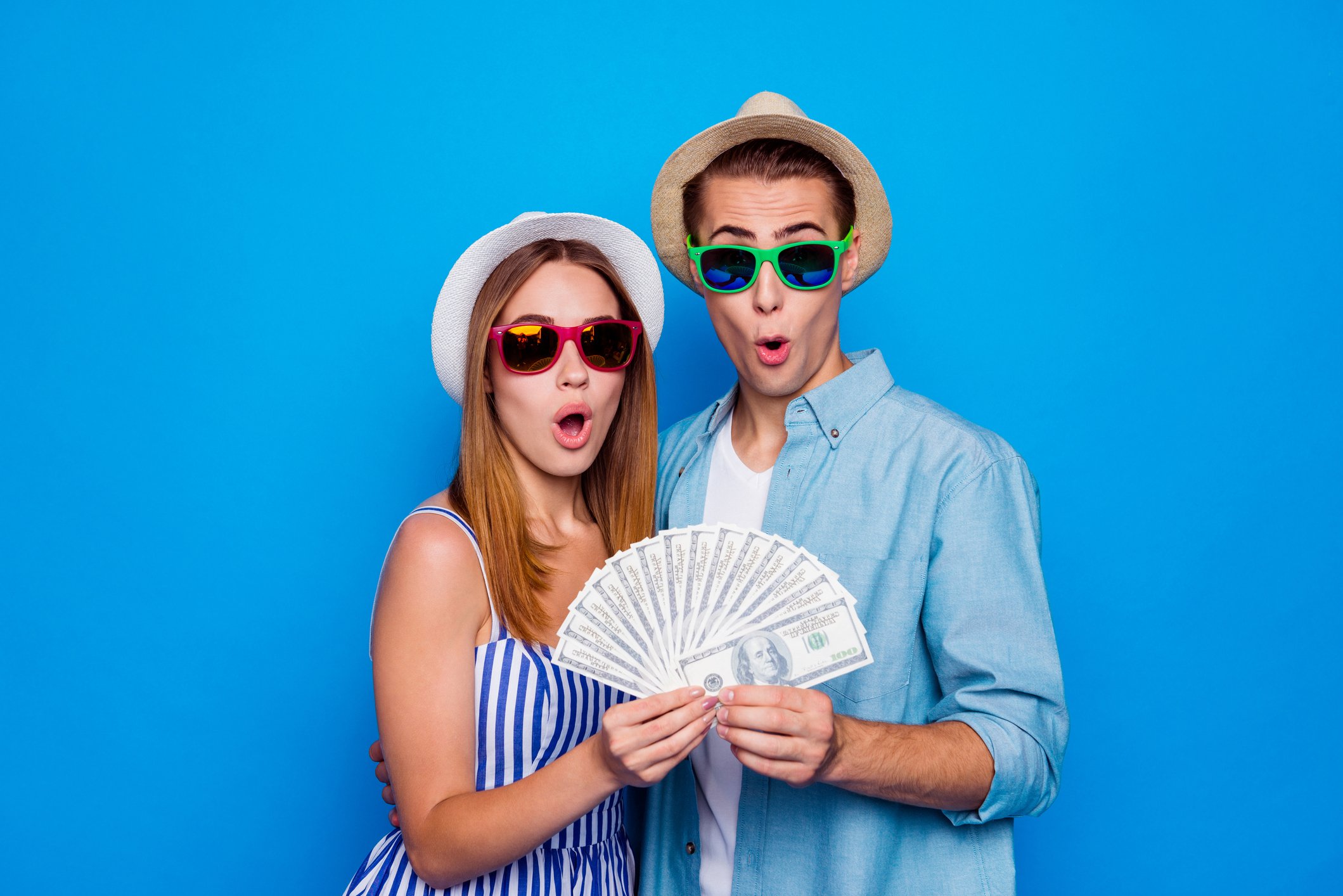 Two cheerfully amazed people in cap holding in hands large sum of money isolated on blue color background