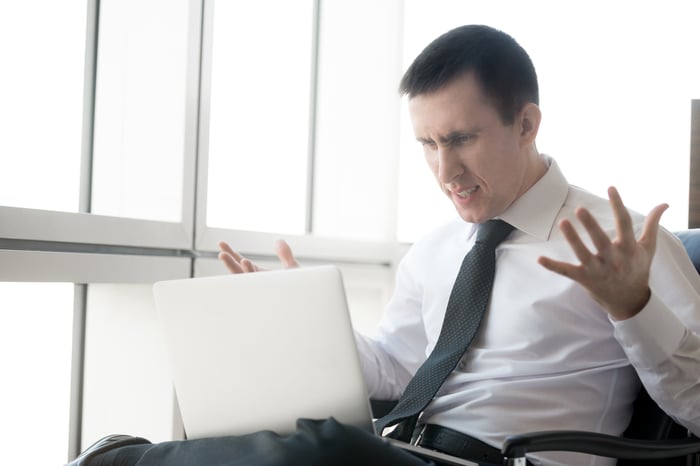 A visibly frustrated businessperson with their hands in the air while looking at an open laptop.