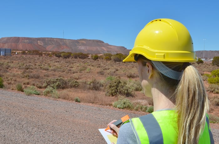 A woman wearing a protective helmet looking over field with pen and paper.