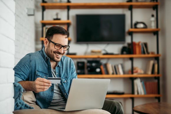 A 30-something man smiles and holds a credit  card while on his laptop in his living room.