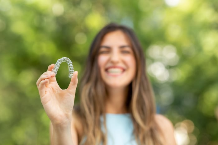 Smiling woman blurred in background holding up a clear invisaligner.