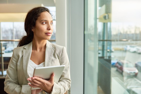A woman dressed in business attire holding a tablet and looking out a window.