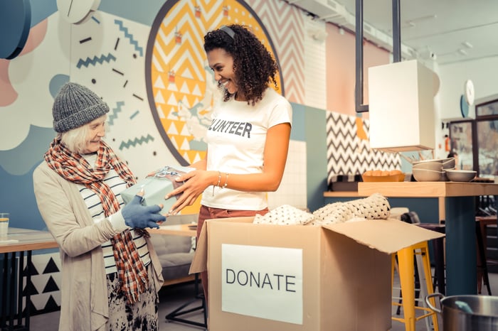 Charity volunteer handing wrapped box to elder woman as they stand next to a donation box.