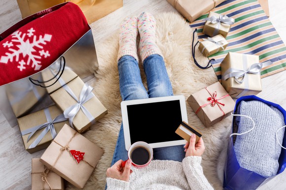 A woman, surrounded by gifts, holds a cup of coffee and shops online in her living room.