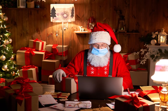 Santa Claus wearing a face covering typing on a laptop surrounded by packages.