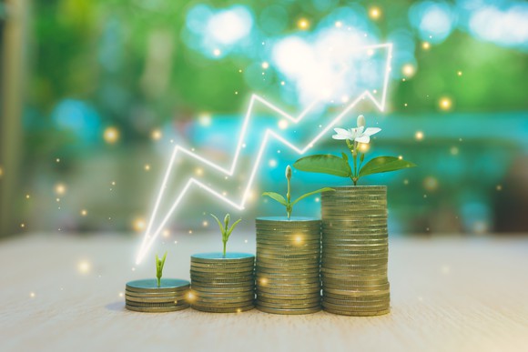 Stacks of coins with plants growing on top are shown on a table witha glowing arrow pointing upward.
