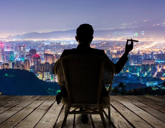 We see the back of a seated man holding a cigar and facing a city lit up at night.