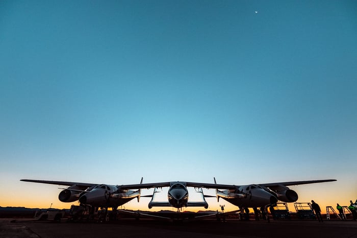 Twin fuselage Virgin Galactic mothership carrying spaceplane at sunrise before test flight.