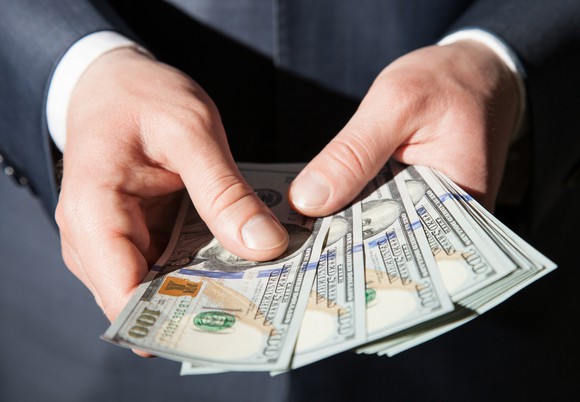 The hands of a man in a suit holding a fanned stack of hundred dollar bills.