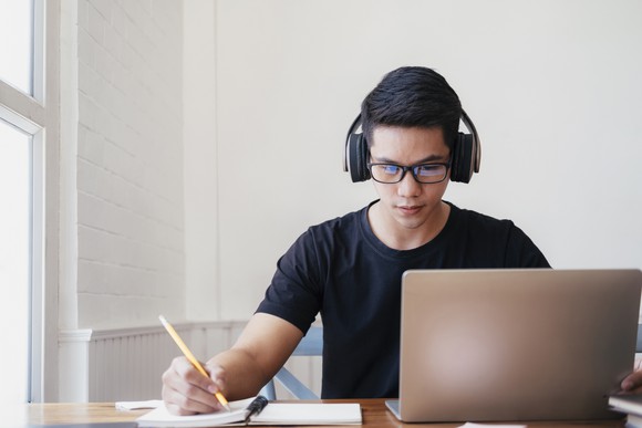 A young person wearing headphones works with a laptop, pencil, and paper.