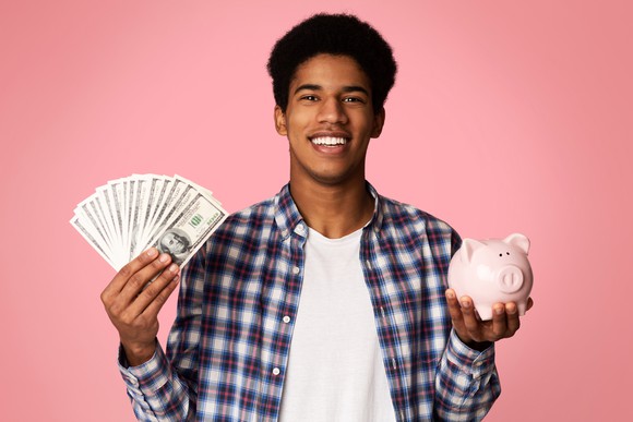 A young man is holding a piggy bank in one hand and cash in the other and smiling.