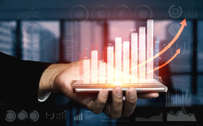 A hand holds a tablet device projecting a holographic image of rising arrows on a bar chart.