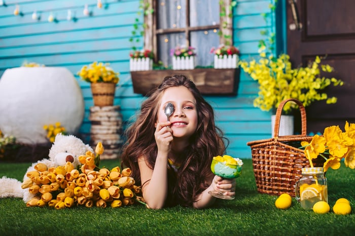 A young girl lying on the lawn in front of a house with a bowl of lemons in her hand and a jar of lemonade nearby.