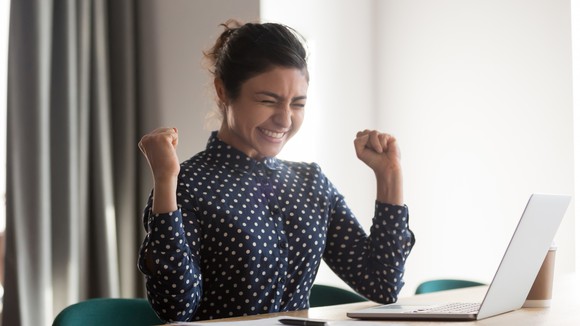 A woman at a desk with her laptop open, raising her fists and smiling.