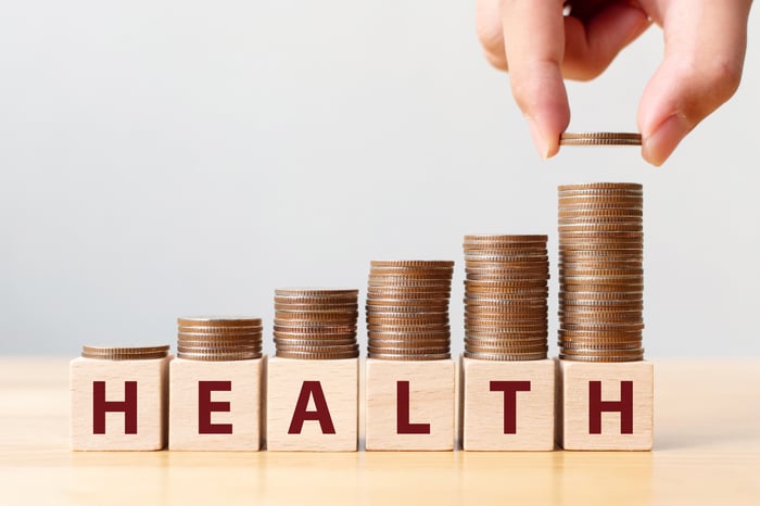 An investor's hand places coins on top of blocks spelling "health."