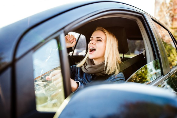 A young woman pumps her fist while singing in the driver's seat of a car.