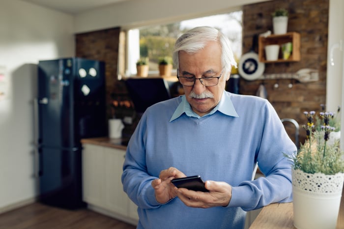 Older man holding mobile phone while standing in the kitchen.