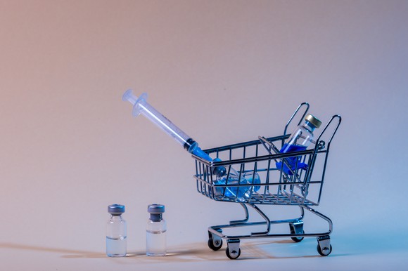 Tiny shopping cart holding a syringe and vaccine vial next to two vaccine vials