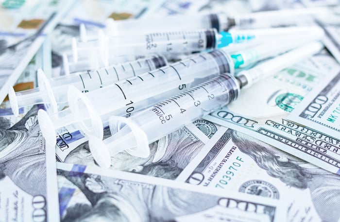 A handful of syringes placed atop a messy pile of one hundred dollar bills. 