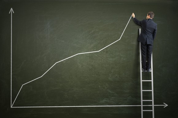 Man on a ladder drawing an upward-bound graph on a board.