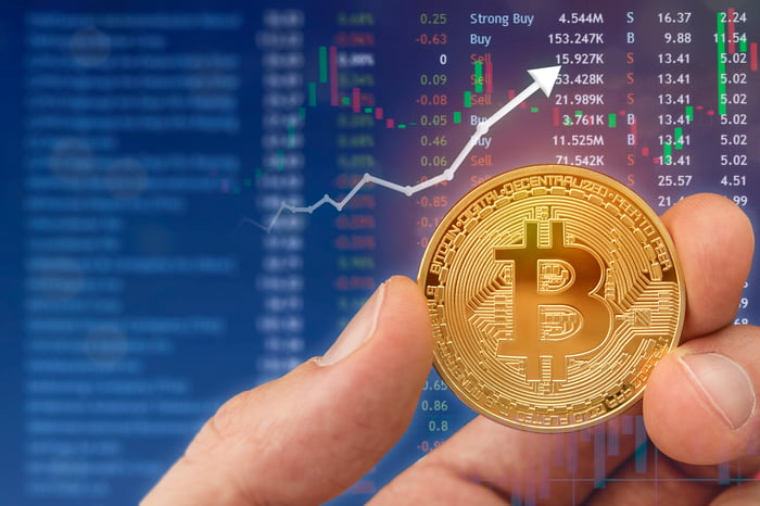 A hand holds a golden coin displaying the Bitcoin symbol with a rising chart in the background.