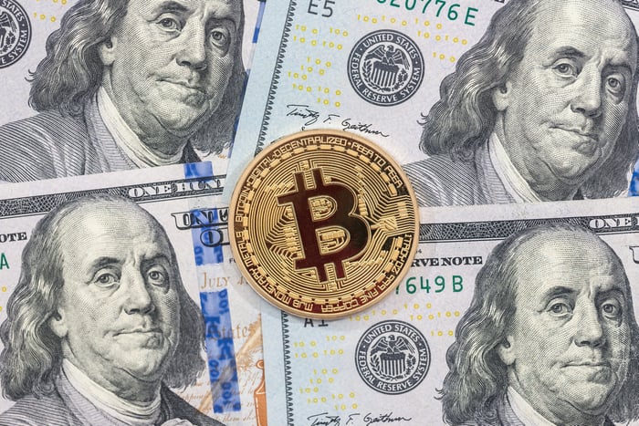 A physical Bitcoin laid atop a pile of one hundred dollar bills.