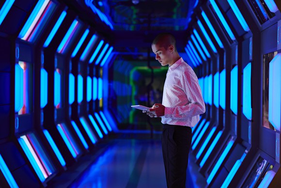 Young man looking down at tablet stands in a spaceship-like corridor.