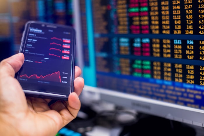 A person holding a smartphone showing stock quotes next to a computer monitor with real-time trade data. 