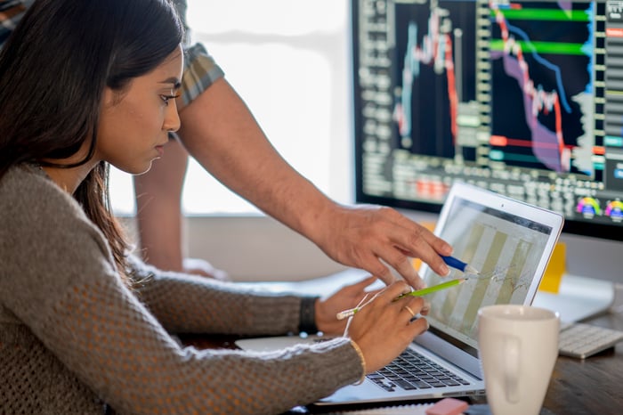 Two people looking at data on a computer with a finance board in the background.