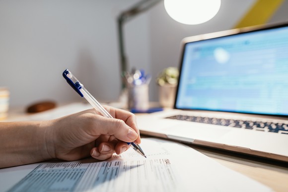 A person filling out a tax form located in front of a laptop on a desk.