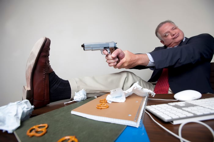 Man in suit sitting at desk aims pistol to shoot himself in the foot