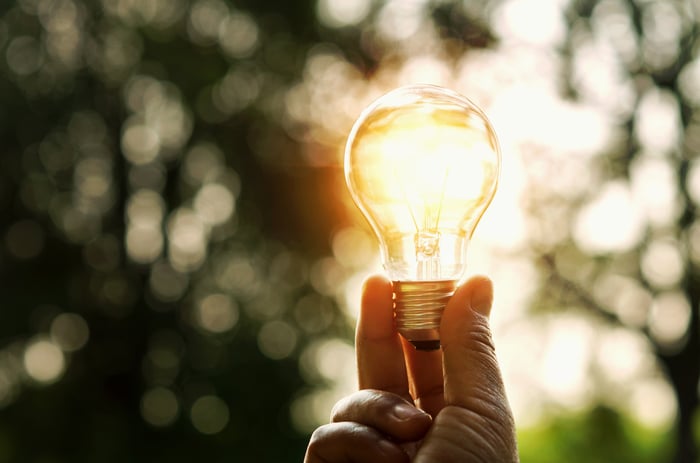 A hand holding up a lit light bulb while outdoors. 