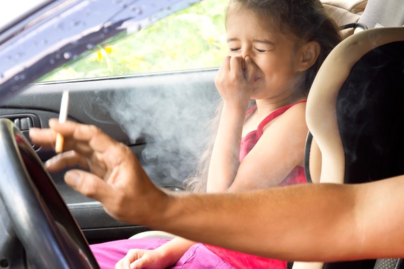 Girl holding her nose in car while adult driver smokes.