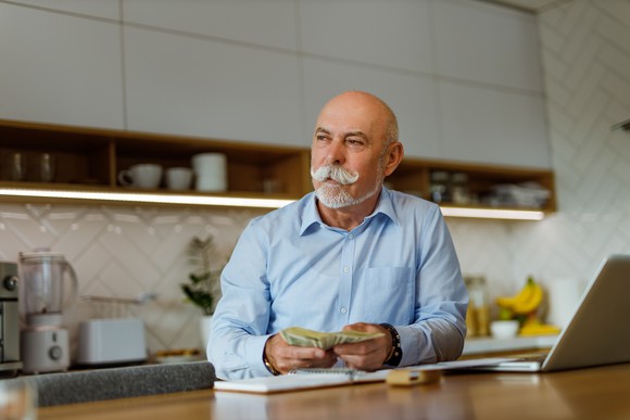 An older well-dressed man sitting in his kitchen holding cash money.