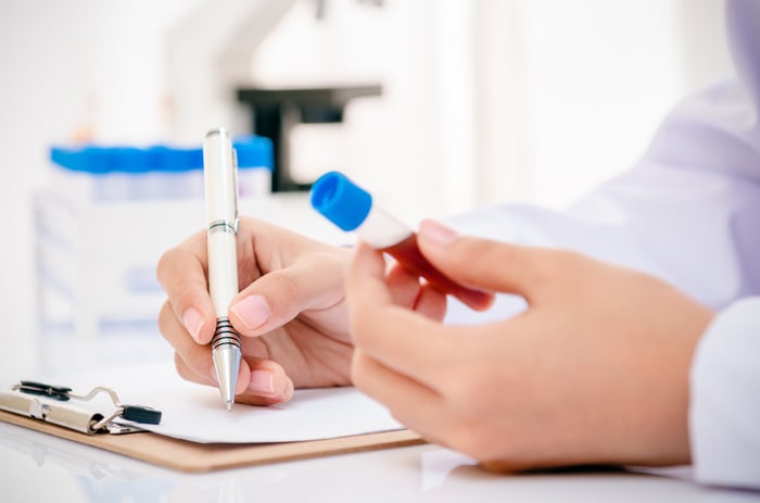 A lab researcher holding a vial of blood in their left hand, while making notes on a clipboard with their right hand.