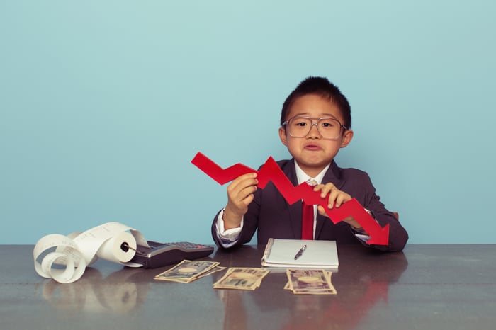 Sitting at table with money on it, a boy holds a down trending red arrow.