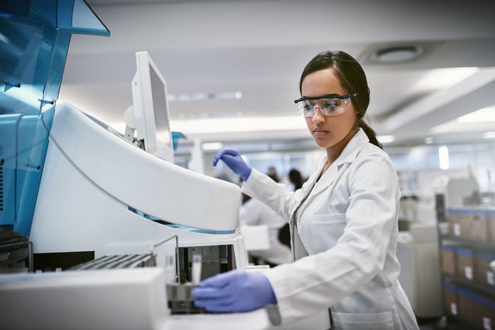 Young woman wearing personal protective equipment and a white coat works at a large machine in a lab.