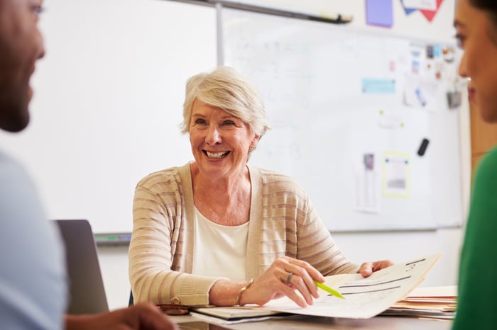 A senior woman sitting at a desk while smiling and holding a sheet of paper.