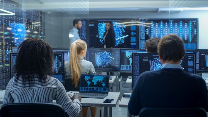 A team of computer technicians in a room with a bank of monitors working on artificial intelligence. 