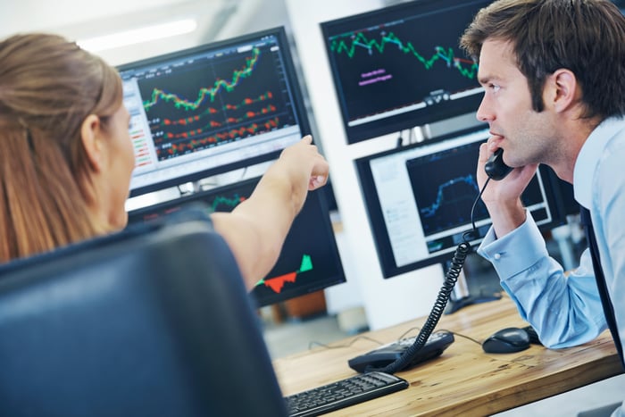 Two professional traders looking at computer monitors featuring charts and investing data. 