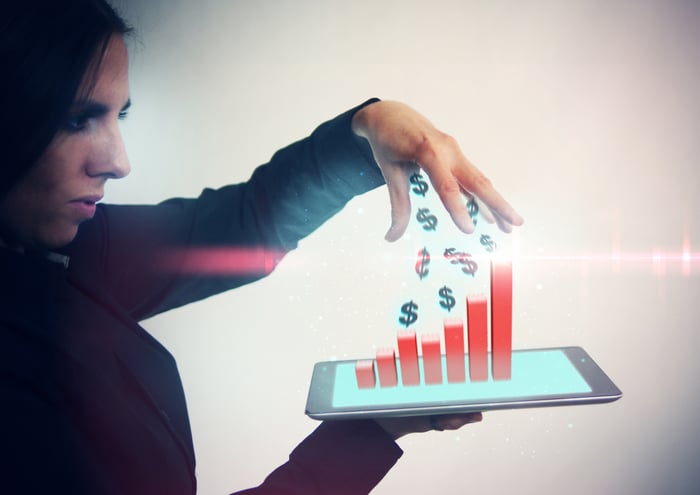 A woman holds a tablet that has a bar graph and dollar signs coming out of it.