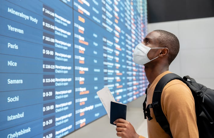 A man wearing a mask, holding his passport and airplane ticket, looking for his flight on the digital board at the airport 