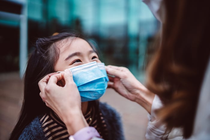 Adult woman places a blue surgical mask over a young girl's nose and mouth.