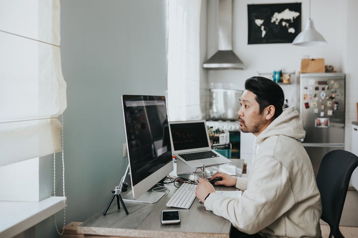 Man wearing a sweatshirt sits at a desk at home in front of a large monitor and laptop.